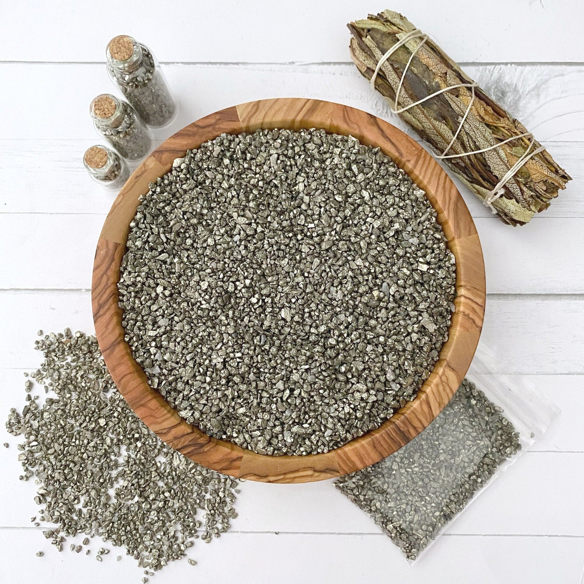 A wooden bowl filled with Pyrite Crystal Chips sits on a white surface. Beside it are two small glass jars with cork stoppers, a bundled sage stick tied with string, and a plastic bag containing more of the Pyrite Crystal Chips. Raw and unpolished tiny stones are also scattered nearby.