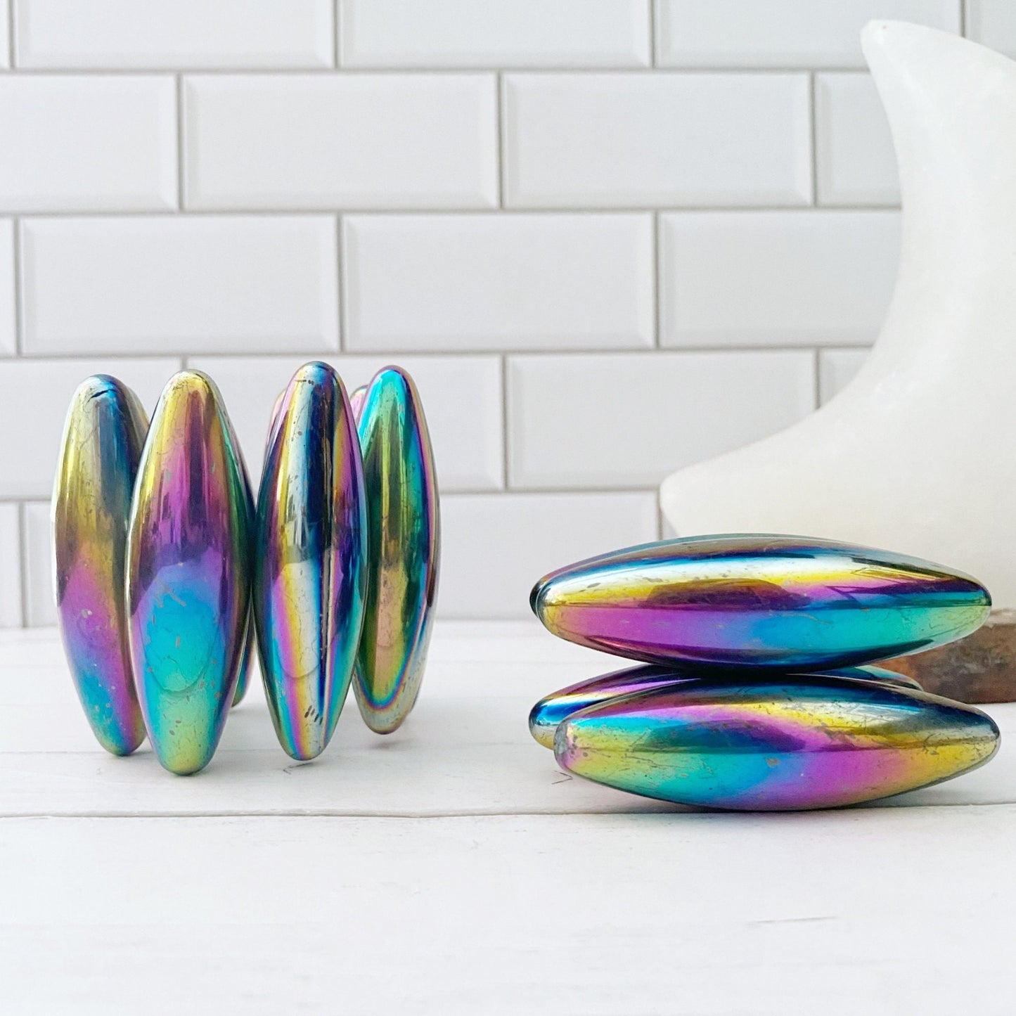 A photo shows two sets of Rainbow Hematite Elliptical Zingers with a lustrous, multi-colored finish. The left set is arranged in a rectangular stack, while the right set forms a pyramid shape. The background features white subway tile and a white geometric object, adding a modern touch to these master grounding stones.