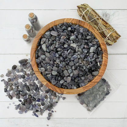 A wooden bowl filled with small, polished purple stones sits on a white surface. Beside the bowl, some tumbled stones are scattered, and a clear bag of Iolite Crystal Chips lays nearby. Three small glass vials containing more stones and a bundle of sage wrapped with string are also present.