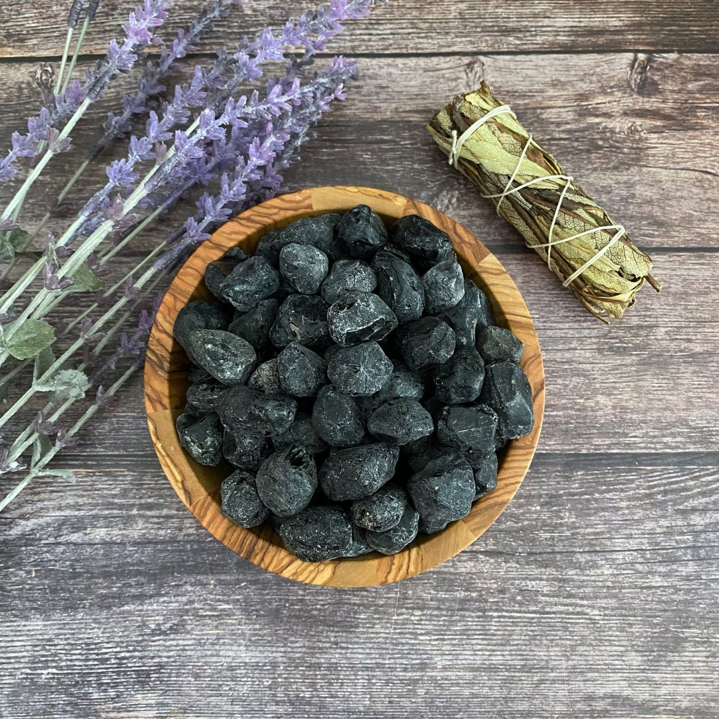 A wooden bowl filled with dark, irregularly shaped Raw Apache Tears sits on a wooden surface. To the left of the bowl are sprigs of lavender, while a bundle of sage wrapped with a string lies just above, offering grounding and grief relief.