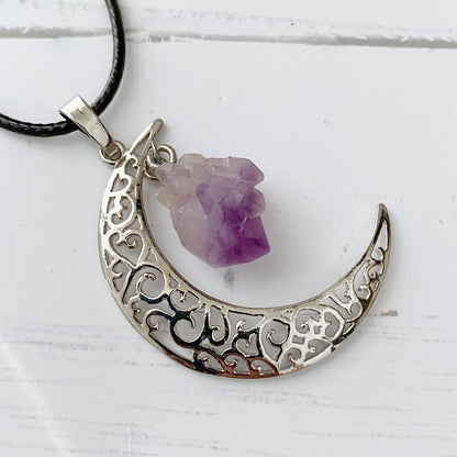 A **Silver Crescent Moon & Amethyst Pendant** with intricate swirls and patterns. Attached to the bottom inside curve is an Amethyst point, known as a protective stone. The pendant hangs on a black cord against a softly blurred gray background.