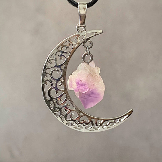 A **Silver Crescent Moon & Amethyst Pendant** with intricate swirls and patterns. Attached to the bottom inside curve is an Amethyst point, known as a protective stone. The pendant hangs on a black cord against a softly blurred gray background.