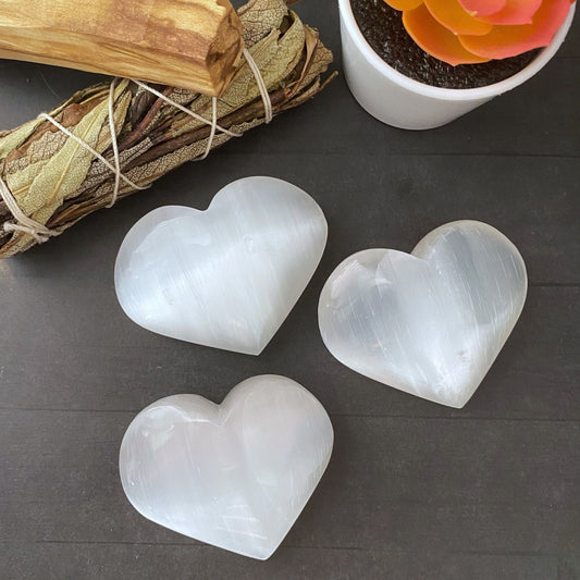 Three polished, translucent 3" Selenite Heart Palm Stones are displayed on a dark surface. Nearby, a bundle of dried sage and a small potted plant with a vibrant orange-red succulent add a touch of natural elements to the arrangement, enhancing the ambiance for spiritual growth.