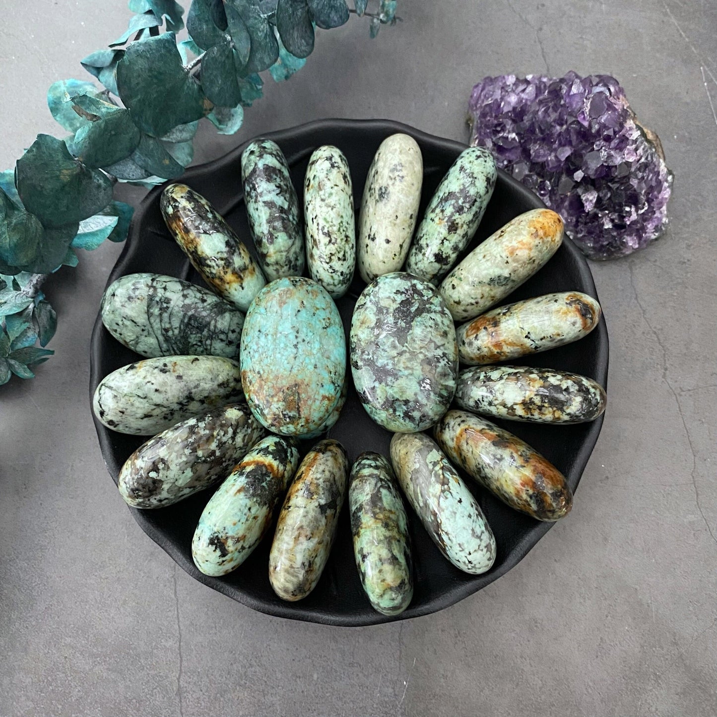 A circular arrangement of polished green and black African Turquoise Palm Stones surrounding a single larger stone in the center, displayed on a dark plate. The background also features a purple amethyst cluster and sprigs of green leaves. The surface is a gray stone texture, enhancing the self-development theme.