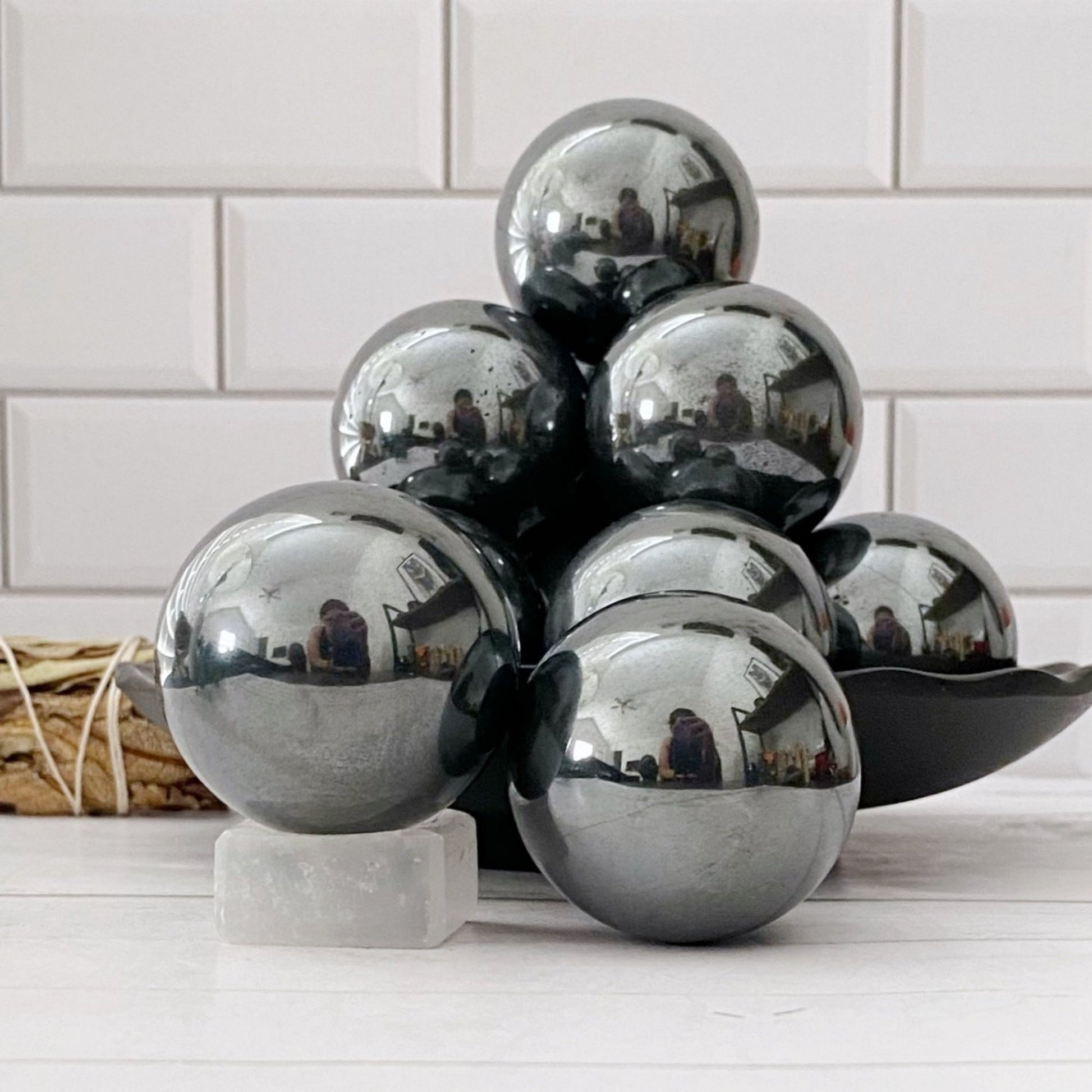 A cluster of reflective metallic balls, likely 40mm Hematite Spheres with their grounding properties, is displayed against a white subway tile background. The spheres are arranged on a dish, capturing reflections of the room and a person on their polished surfaces. A small white cube sits in the foreground.