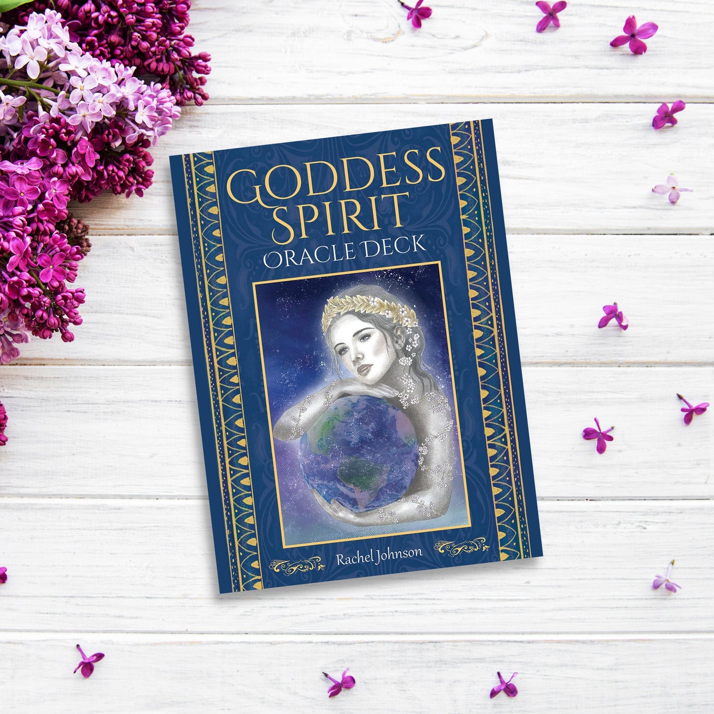 A blue book titled "Goddess Spirit Oracle" by Rachel Johnson rests on a white wooden surface, surrounded by purple flowers. The cover, embracing mystical femininity, features an illustration of a woman holding a globe, adorned with a celestial and ethereal aesthetic.