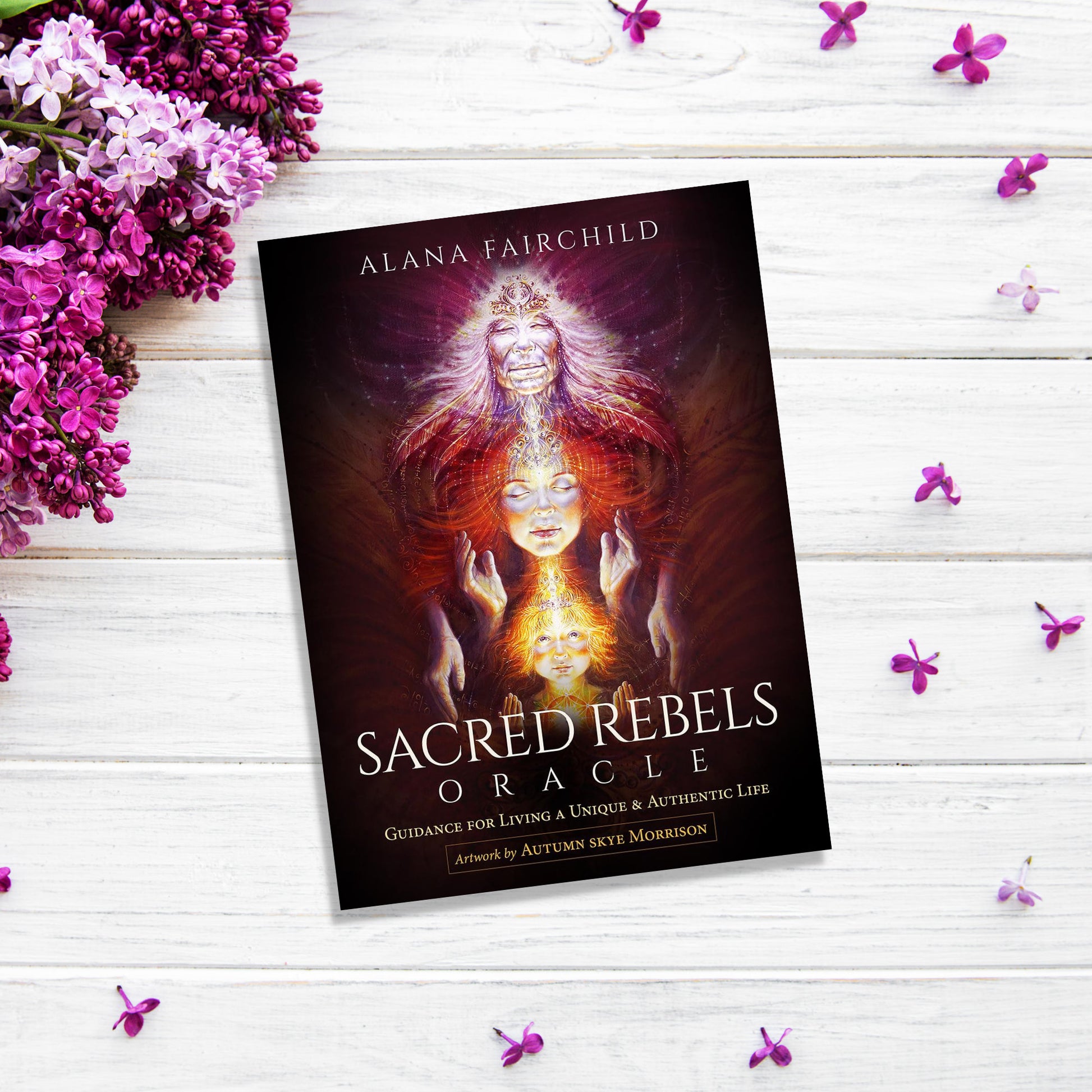 A book titled "Sacred Rebels Oracle" by Alana Fairchild lies on a white wooden surface. The cover features mystical artwork of radiant figures manifesting dreams. Purple flowers frame the image, adding a vibrant touch.