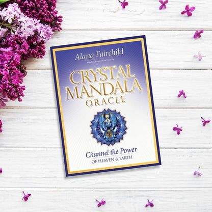 The image shows the cover of the "Crystal Mandala Oracle" by Alana Fairchild placed on a white wooden surface, surrounded by pink and purple flowers. The cover features a mandala design in the center with a gradient purple and yellow background, radiating divine healing power.