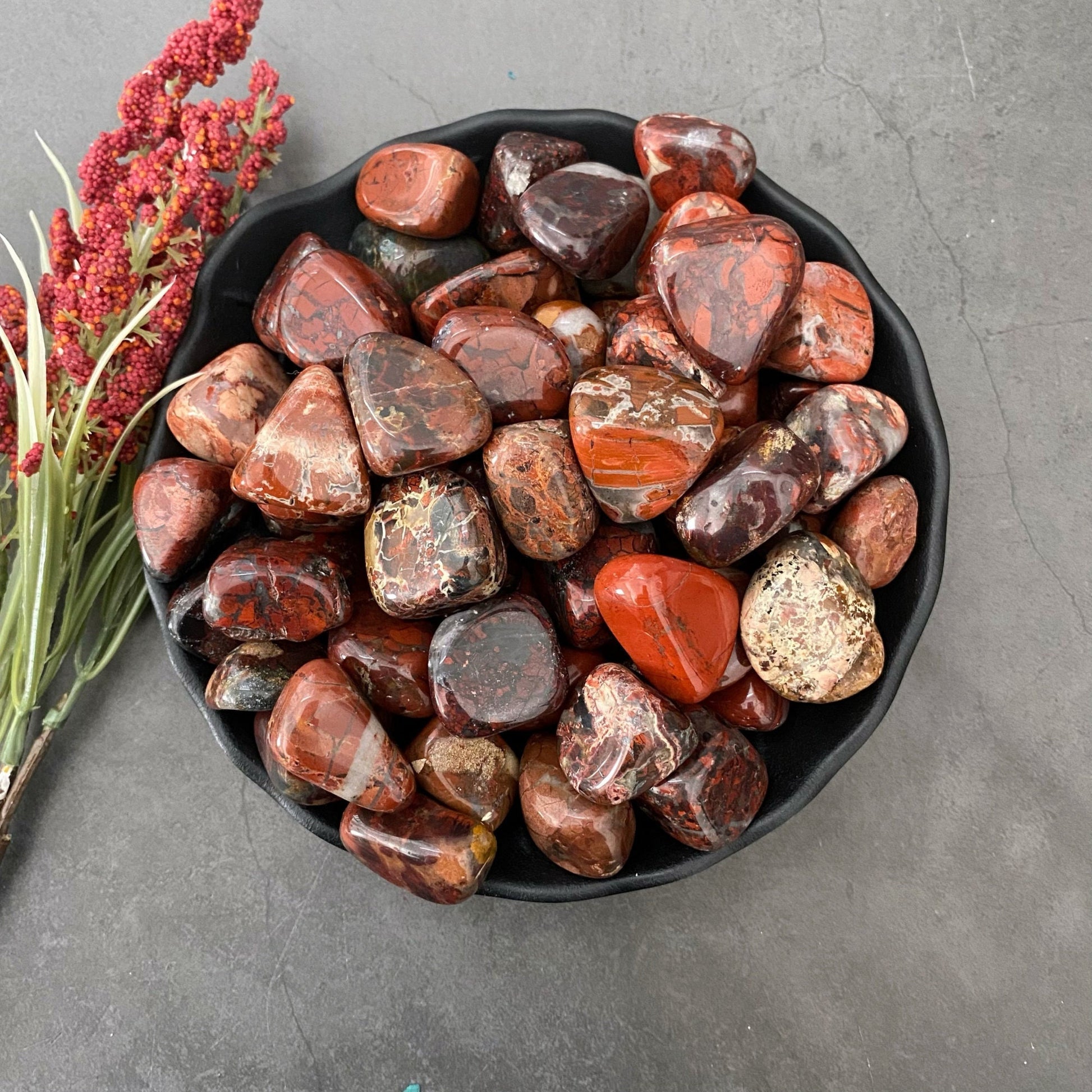 A black bowl filled with polished, multicolored jasper stones, predominantly in earthy tones such as reds, browns, and oranges, sits on a gray surface. The Brecciated Jasper Tumbled Stones are known for their grounding energy. To the left of the bowl, there is a small bunch of red, dried flowers.