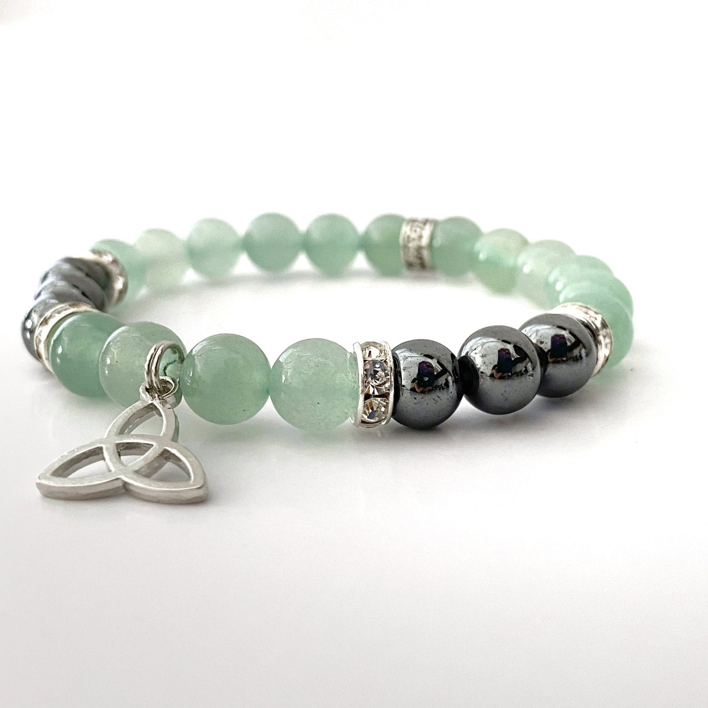 An 8mm Green Aventurine & Hematite Beaded Bracelet featuring light green beads interspersed with dark gray beads and silver accents. At the forefront is a silver Triquetra charm with an intricate, triangular knot design. The bracelet is displayed on a clean, white background.