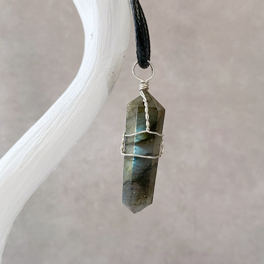 A 1.5" Double Terminated Labradorite Point Pendant wrapped in silver wire, attached to a black cord, is displayed against a soft gray background. The crystal has iridescent shades of blue and green and hangs elegantly from a white antler-shaped holder.