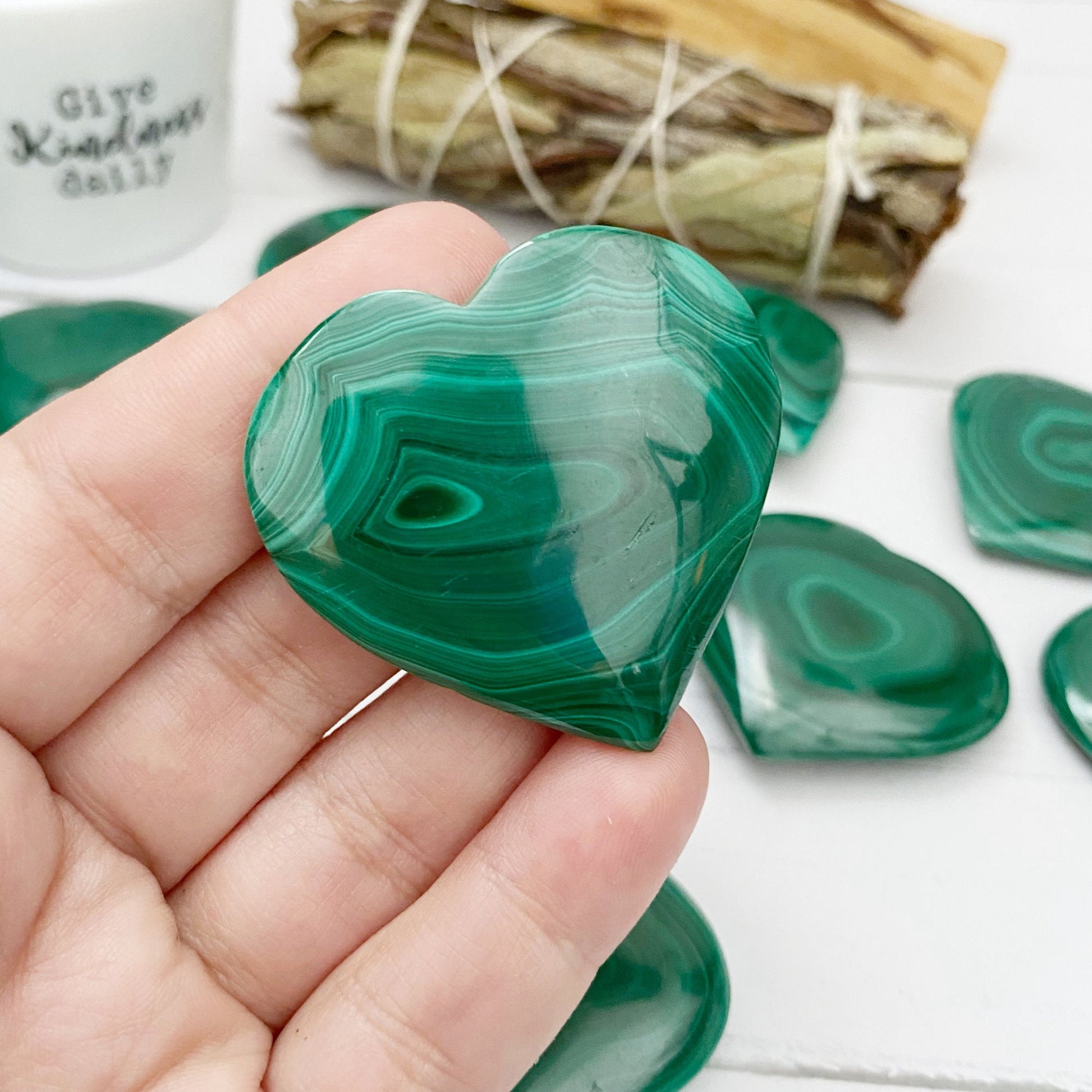 A collection of 1.5" Small Malachite Hearts with swirling patterns is neatly arranged on a white wooden surface. Off to the side, a plant in a white pot and a bundle of tied wooden sticks are visible, adding to the aesthetic. Known as protection stones, these malachite pieces absorb negative energies beautifully.