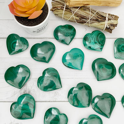 A collection of 1.5" Small Malachite Hearts with swirling patterns is neatly arranged on a white wooden surface. Off to the side, a plant in a white pot and a bundle of tied wooden sticks are visible, adding to the aesthetic. Known as protection stones, these malachite pieces absorb negative energies beautifully.