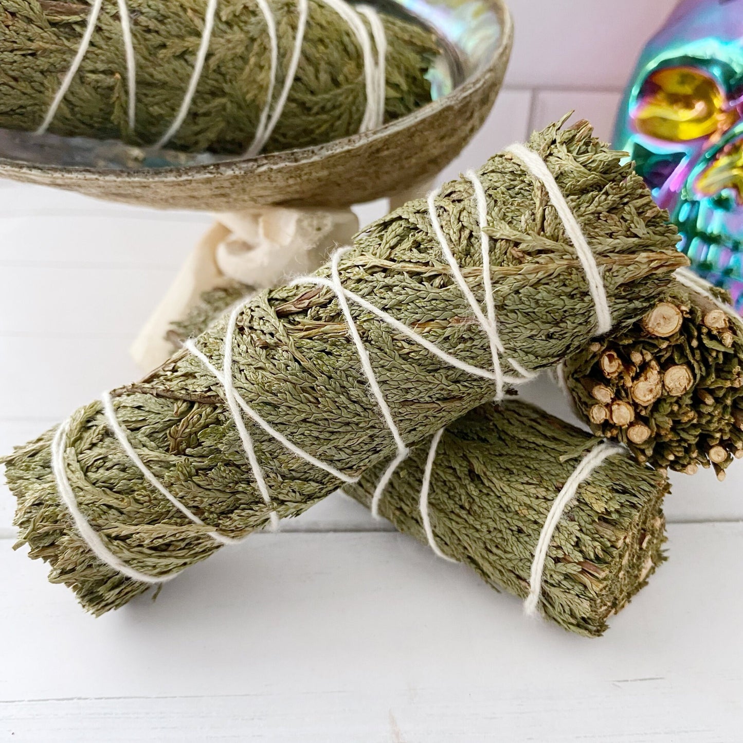 Three bundles of Organic Cedar Sage Smudge Stick, ideal for energy cleansing, are wrapped in white string and displayed on a white surface. One bundle rests on a seashell, while the others lie in front. In the background, a partially blurred colorful container with a plant adds a pop of color against the white subway tile backdrop.