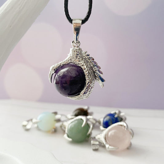 A close-up of a Dragon Claw Gemstone Pendant featuring an amethyst gemstone sphere held in a silver claw setting on a black cord. Blurred in the background are other similar pendants with different colored gemstone spheres, including blue, green, pink, and white.