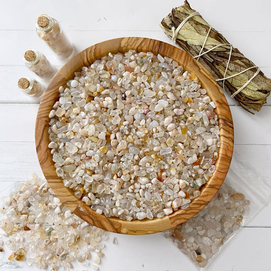 A wooden bowl filled with small, polished Flower Agate Crystal Chips on a white surface. Three small glass jars with cork stoppers containing similar crystals are positioned nearby. A bundle of dried sage leaves is tied with a string, and a small bag of crystals is partially opened, showcasing their metaphysical uses.