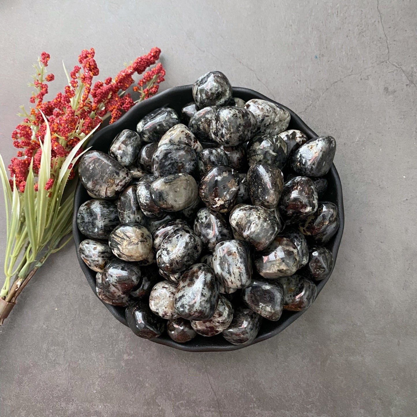 A black bowl filled with polished black and white stones, including Astrophyllite Tumbled Stones, is placed on a gray surface. To the left of the bowl, there is a small arrangement of red berries and green leaves. The overall setup has a natural and earthy aesthetic, perfect for chakra realignment.