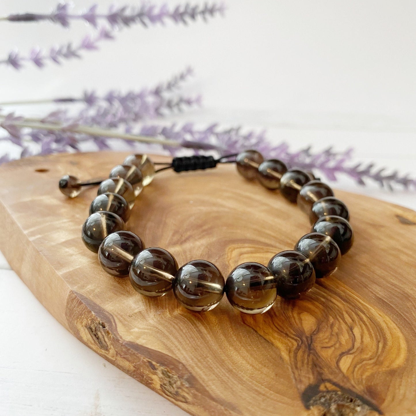 A 10mm Adjustable Smoky Quartz Beaded Bracelet featuring glossy, round, dark brown translucent beads made from grounding Smoky Quartz is displayed against a plain white background. The Reiki energy-infused beads are strung together on an adjustable elastic cord with a black knot visible in the background.