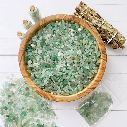 A wooden bowl filled with green and white sea glass pieces sits on a white surface, scattered with tiny Green Aventurine Crystal Chips. Some sea glass pieces are scattered outside the bowl. Small glass bottles with sea glass and a bundle of tied sticks are also present in the background.