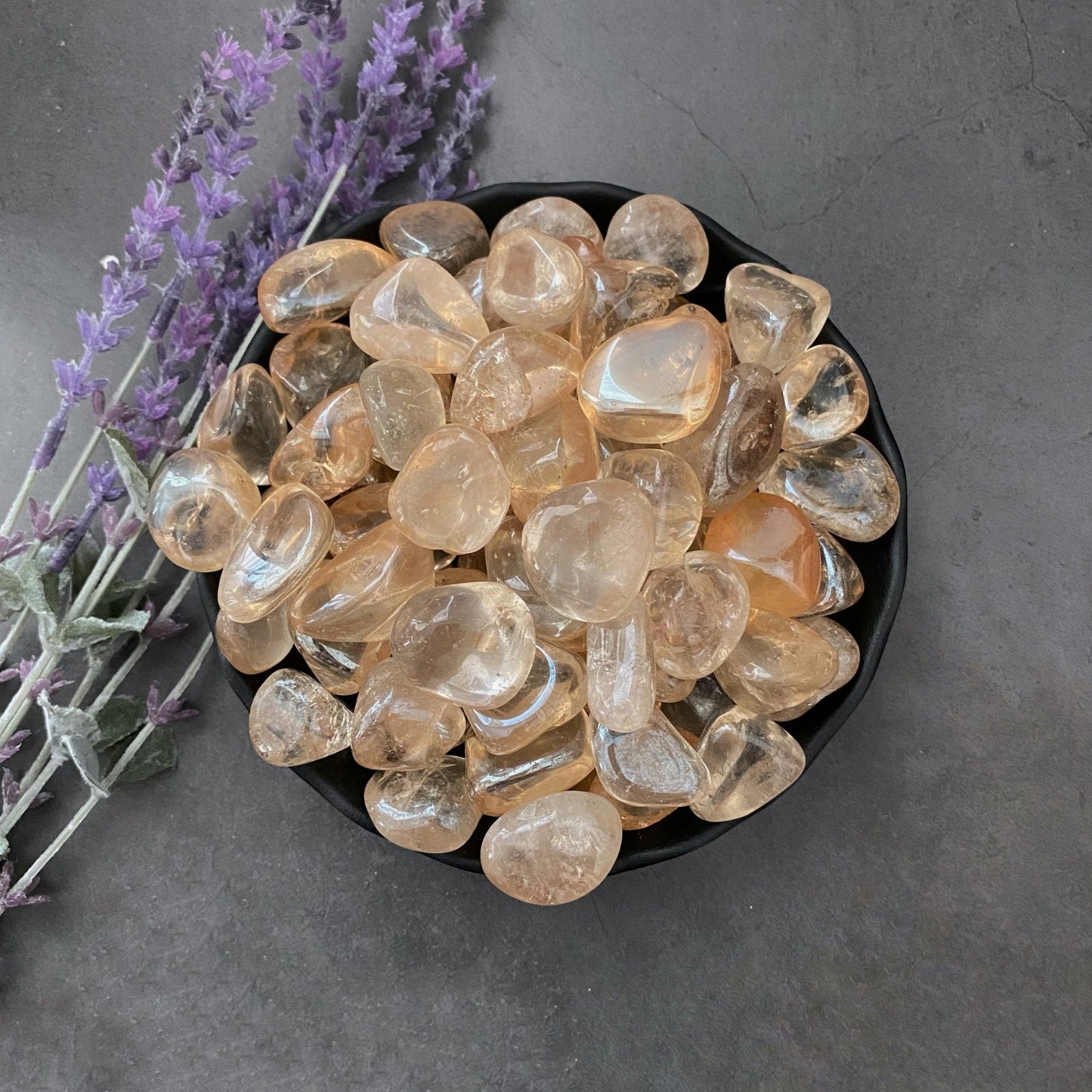 A black bowl filled with polished, translucent Champagne Aura Quartz Tumbled Stone—an abundance crystal prized for its energy—is placed on a dark, textured surface. Beside the bowl, a few sprigs of purple lavender flowers lie, adding a touch of color to the composition.