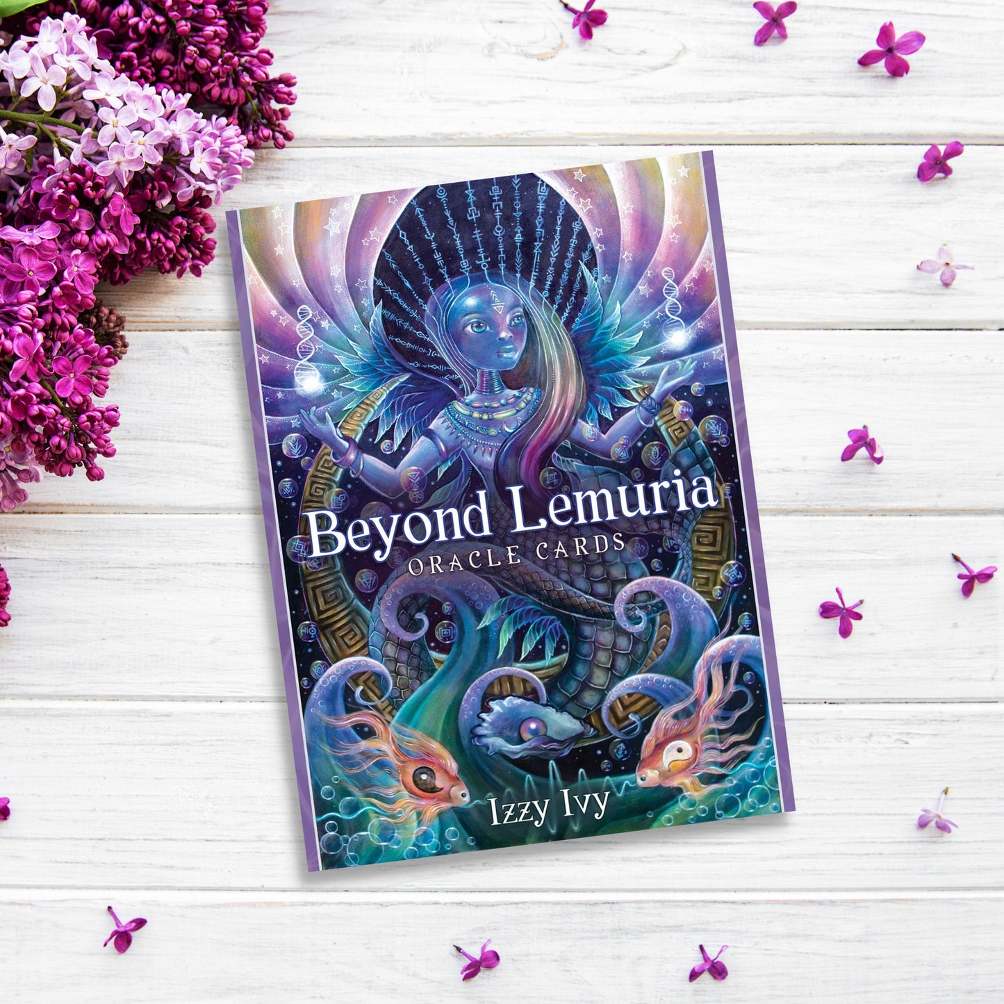 A deck of Beyond Lemuria Oracle Cards by Izzy Ivy is placed on a white wooden surface surrounded by purple flowers. The cover features a mystical being with an elaborate headdress and aquatic elements, including fish and tentacles, set against a vibrant, colorful background that evokes conscious awareness.