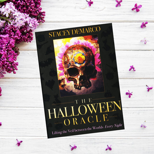 A book titled "The Halloween Oracle" by Stacey Demarco lies on a white wooden surface, surrounded by purple flowers. The cover features a skull with intricate colorful designs, including flowers and a clock, set against a vivid background—perfect for those interested in divination.