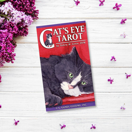 A tarot deck titled "Cat's Eye Tarot Card Deck" by Debra M. Givin, DVM is placed on a white wooden surface, surrounded by purple flowers. The cover features an illustration of a black and white cat with green eyes lying down, looking at the viewer—inspired by Rider-Waite interpretations.