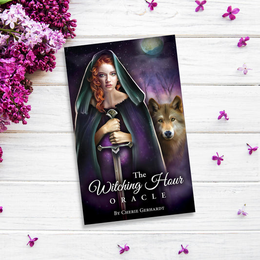 A book titled "The Witching Hour Oracle" by Cherie Gerhardt lying on a white wooden surface. The cover art depicts a red-haired woman in a dark cloak holding a sword, with a wolf and a moonlit night sky in the background. Surrounding "The Witching Hour Oracle" and its Triple Goddess energy are purple flowers.