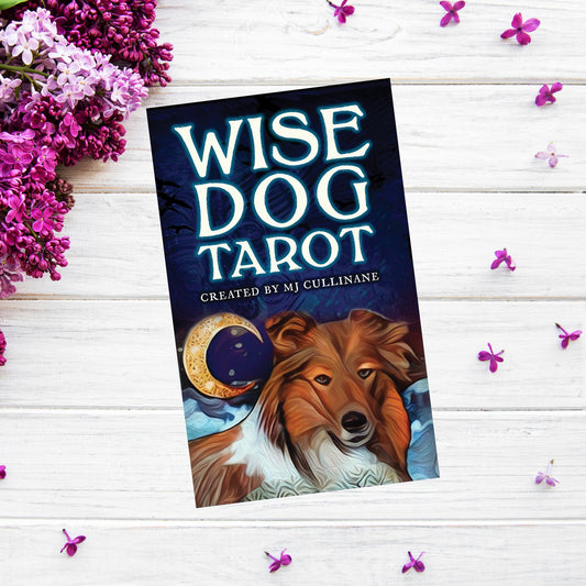 A tarot card deck titled Wise Dog Tarot by MJ Cullinane is positioned on a wooden surface surrounded by purple flowers. The cover features a thoughtful-looking dog with a crescent moon in the background, emphasizing rich dog symbolism throughout the deck.