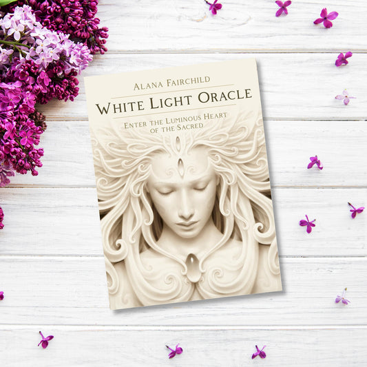 A product titled "White Light Oracle" by Alana Fairchild lies on a white wooden surface, surrounded by purple flowers. The cover, featuring an ethereal figure with flowing hair and intricate designs, offers divine medicine for the soul.