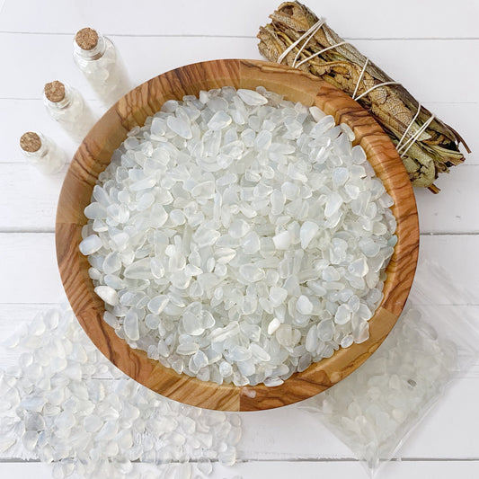 A wooden bowl filled with translucent Opalite Crystal Chips is placed on a white wooden surface. Beside it, there are small bundles of sage, tumbled and polished stones, and three small glass bottles with corks. A plastic bag and scattered Opalite Crystal Chips are also visible around the bowl.