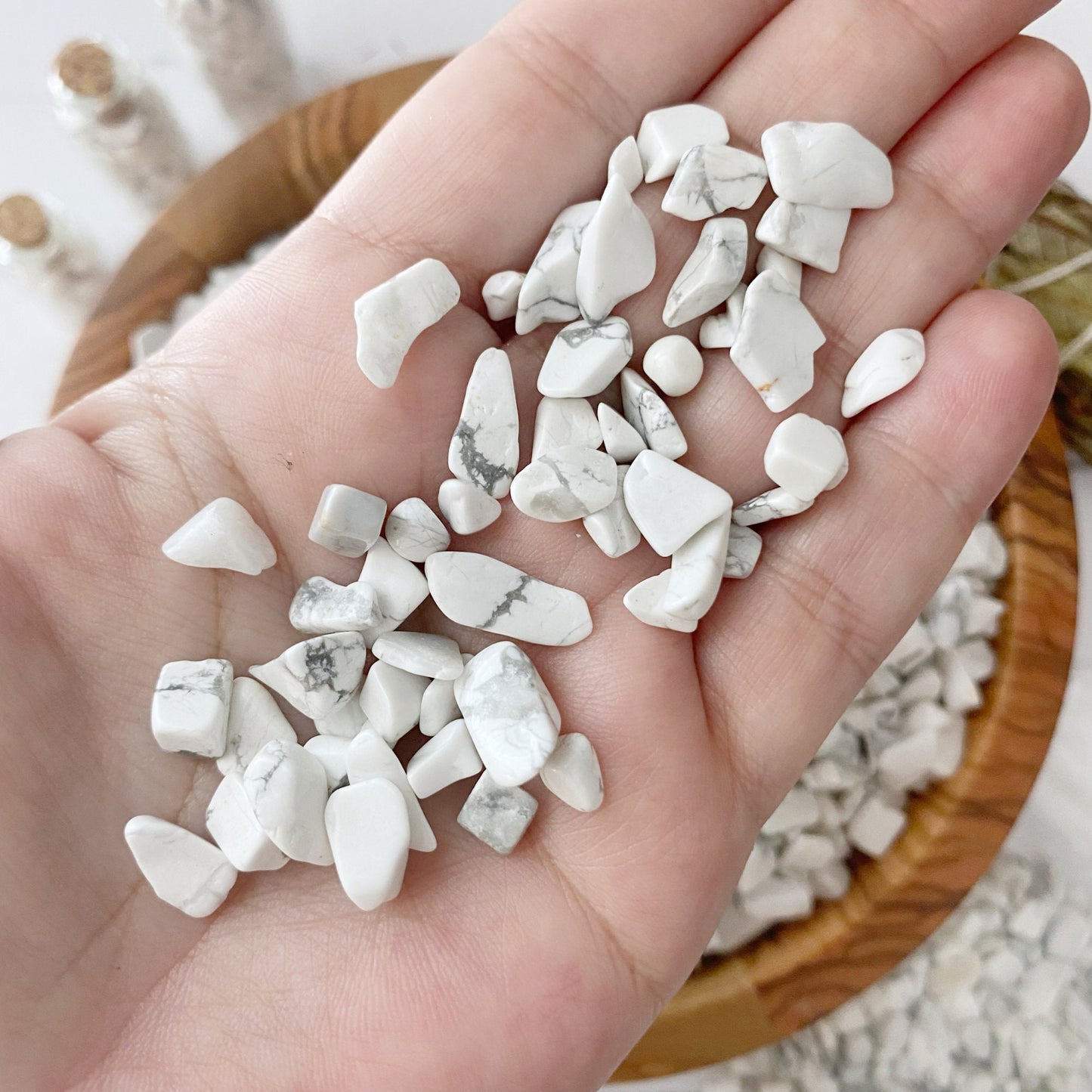 A wooden bowl filled with tumbled and polished stones, including Howlite Crystal Chips, rests on a white surface. Some stones are scattered outside the bowl. Three small glass bottles and a bundle of dried sage sticks are placed beside the bowl, creating a serene setting for emotional healing.