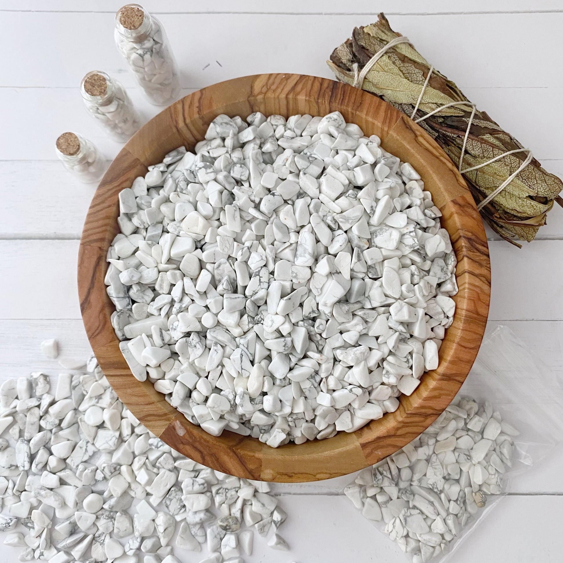 A wooden bowl filled with tumbled and polished stones, including Howlite Crystal Chips, rests on a white surface. Some stones are scattered outside the bowl. Three small glass bottles and a bundle of dried sage sticks are placed beside the bowl, creating a serene setting for emotional healing.