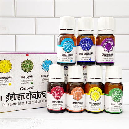 A set of seven therapeutic grade essential oil bottles labeled with different chakras: Root, Sacral, Solar Plexus, Heart, Throat, Third Eye, and Crown. They are arranged in a pyramid shape beside a box labeled "7 Chakra Essential Oil Blends" on a white tiled background.