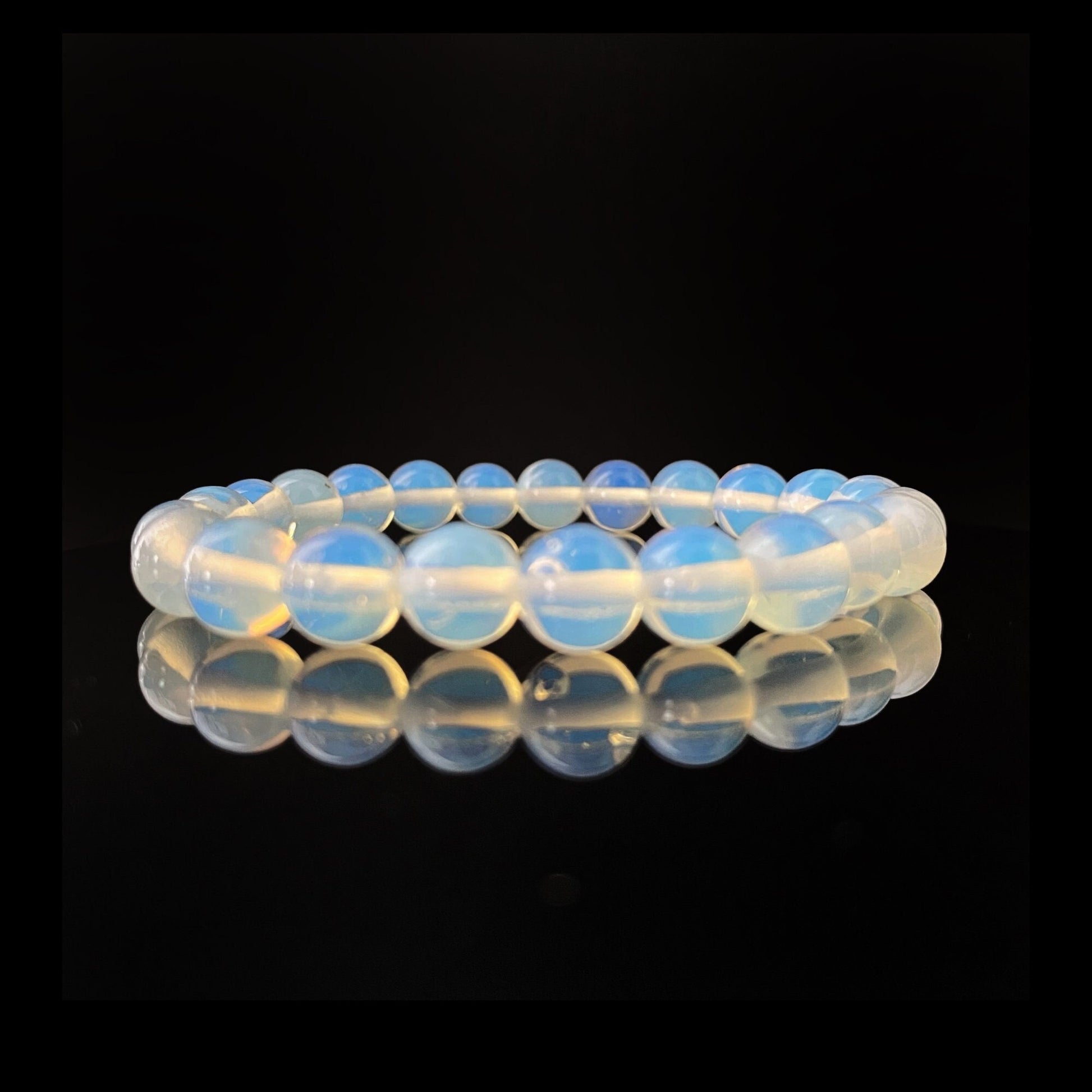 A close-up photograph of an 8mm Opalite Beaded Bracelet against a black background. The spherical, evenly sized beads emit a soft, milky glow with subtle blue and yellow hues. Known as a meditation stone with healing properties, the bracelet's reflection shimmers on the shiny surface below.