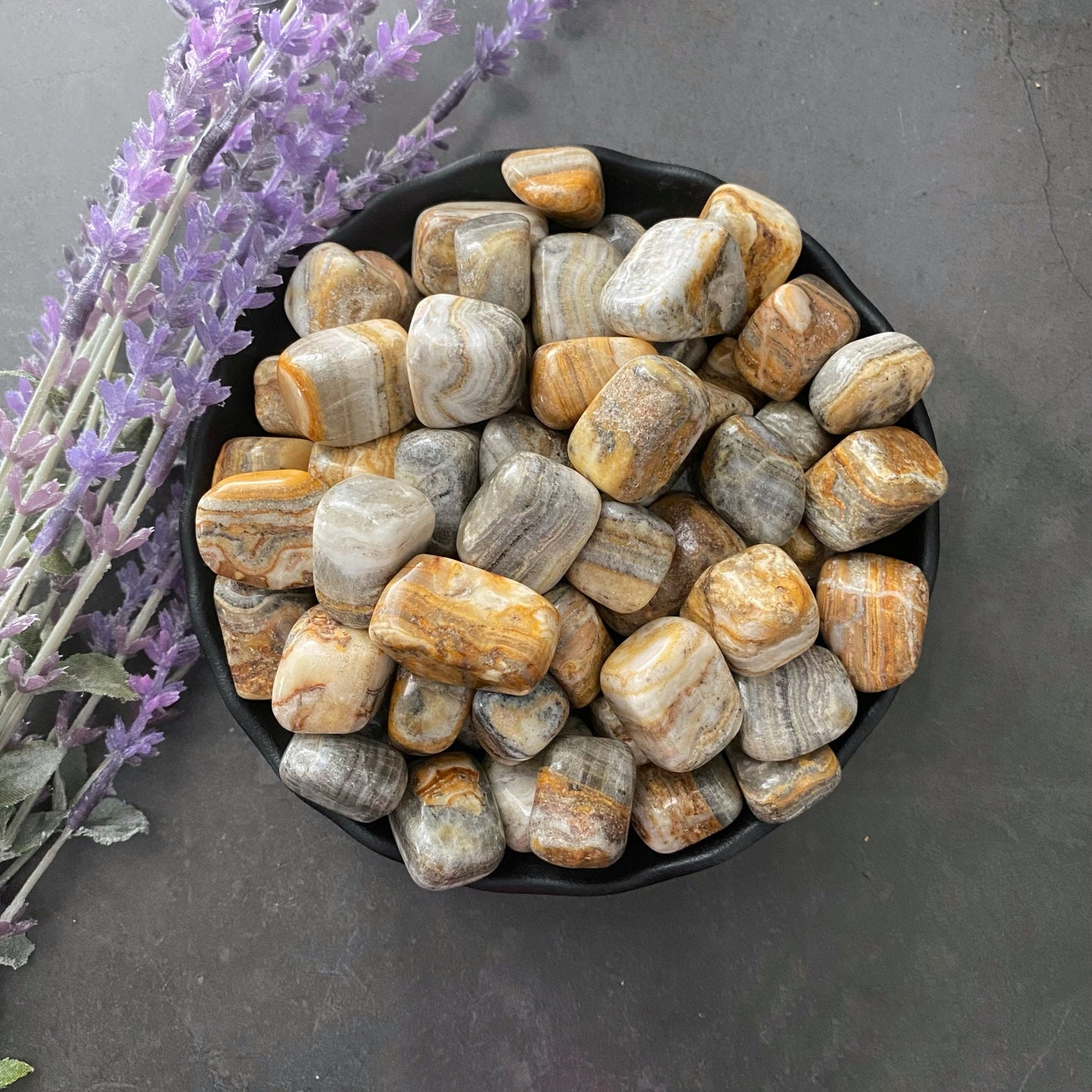 A black bowl filled with numerous Banded Onyx Tumbled Stones, including shades of gray, yellow, and brown, is placed on a black surface. To the left of the bowl, there is a sprig of lavender flowers.