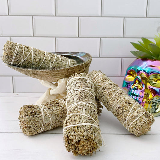 A collection of four 4" Organic Blue & White Sage Smudge Sticks wrapped in white string lies on a white wooden surface. Behind them is an abalone shell supported by a cloth, and to the right, a metallic, rainbow-colored decorative skull. A small succulent is partially visible.