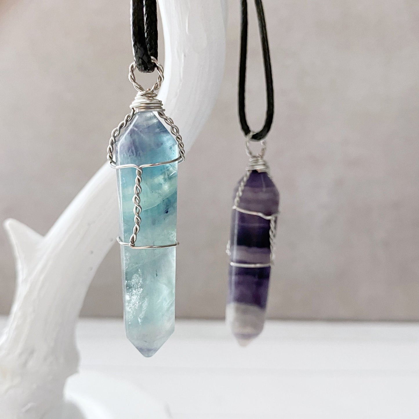 Four 1.5" Double Terminated Rainbow Fluorite Wire Wrapped Pendants hang from black cords against a white background. The crystals, wire-wrapped in silver, display gradient hues of blue, purple, green, and clear, showcasing their natural variations and polished finish.