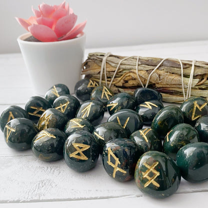 A collection of polished dark green Bloodstone Elder Futhark Runes, featuring gold symbols, is arranged on a white surface. A bundle of sage wrapped with twine and a small white pot holding a pink succulent are placed in the background.