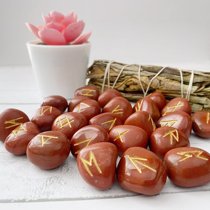 Red Jasper Elder Futhark Runes are laid out on a white wooden surface. Behind them is a bundle of sage tied with string, adjacent to a small potted pink succulent plant. The background is a light grey.