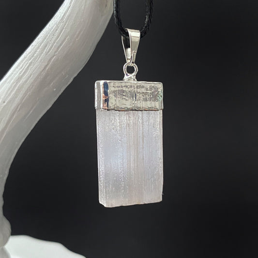 A Raw Selenite Pendant with Silver Cap with purifying properties, enclosed in a silver-finish metal cap, hangs from a black braided cord against a dark background. The textured, shining surface of the rectangular pendant is showcased on a white, curved stand.