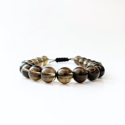 A 10mm Adjustable Smoky Quartz Beaded Bracelet featuring glossy, round, dark brown translucent beads made from grounding Smoky Quartz is displayed against a plain white background. The Reiki energy-infused beads are strung together on an adjustable elastic cord with a black knot visible in the background.