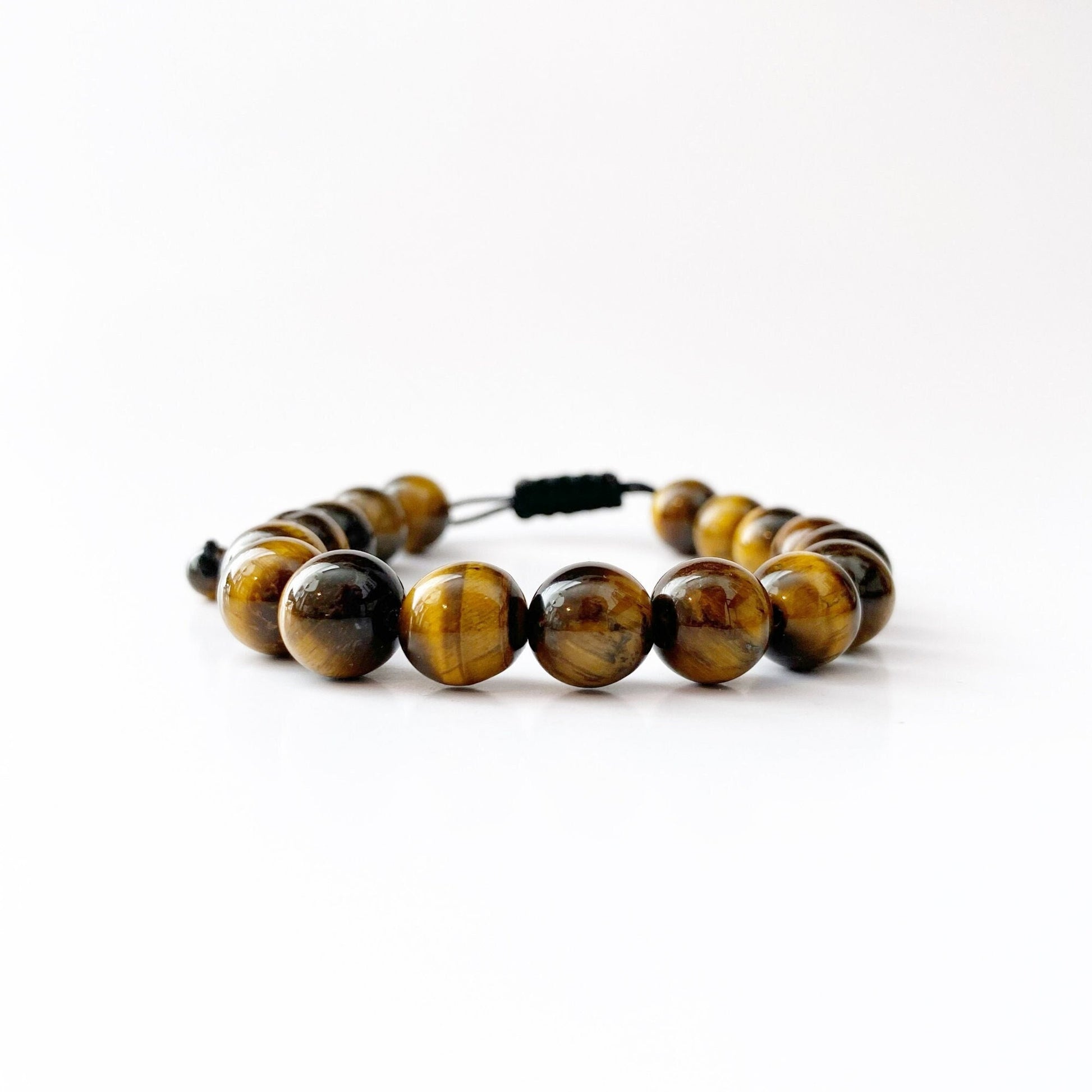 A polished 10mm Tiger Eye Beaded Bracelet with smooth, round beads strung together on a black cord. The beads showcase a blend of golden brown, yellow, and dark brown hues with a reflective sheen, lying on a white background. This beaded bracelet harmonizes the natural beauty of the stone with serene Usui Reiki energy.
