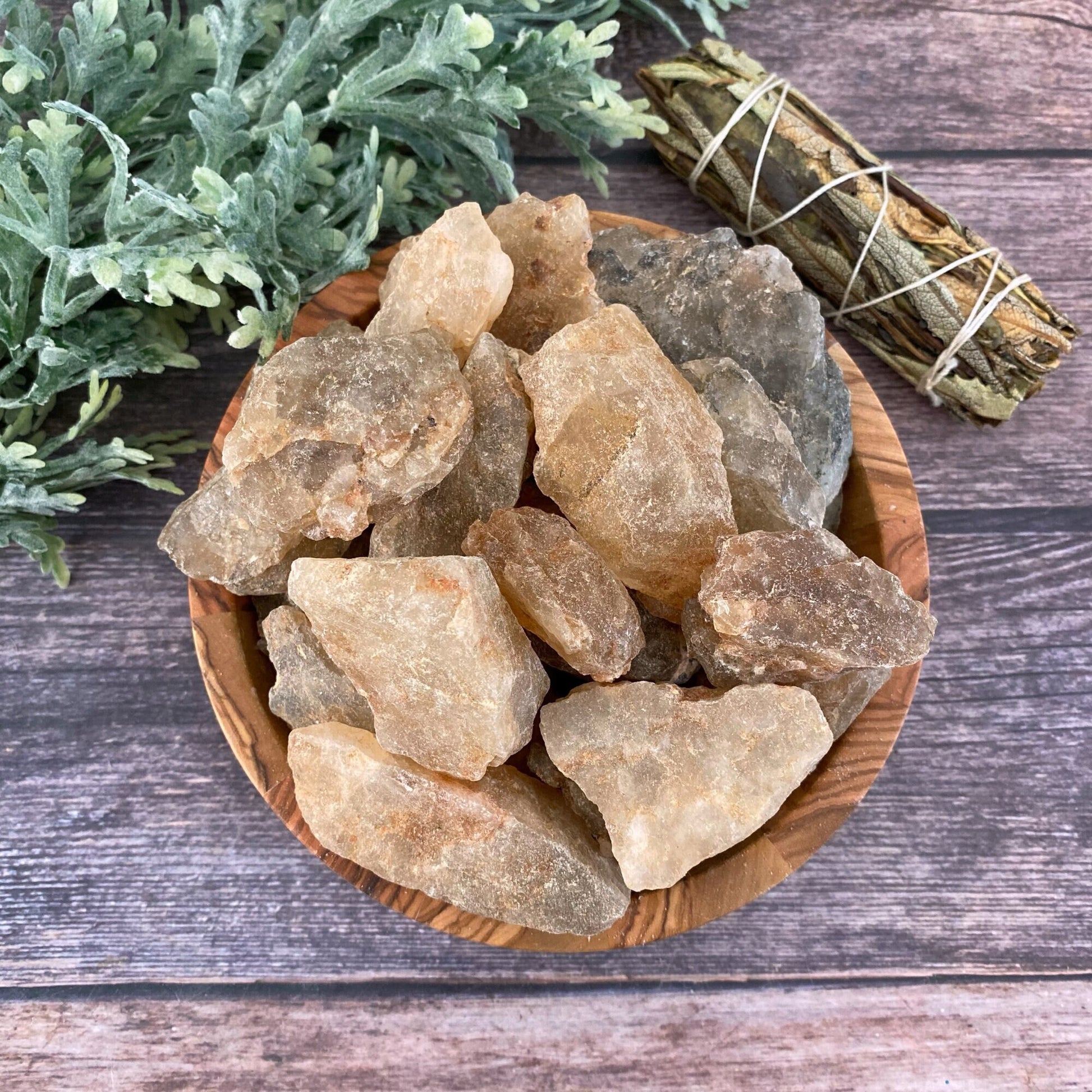 A wooden bowl filled with large, rough chunks of Raw Natural Topaz Stones sits on a wooden surface. Behind the bowl, there is a bundle of sage tied with string and some green leafy foliage, emphasizing the metaphysical properties often associated with the root chakra.