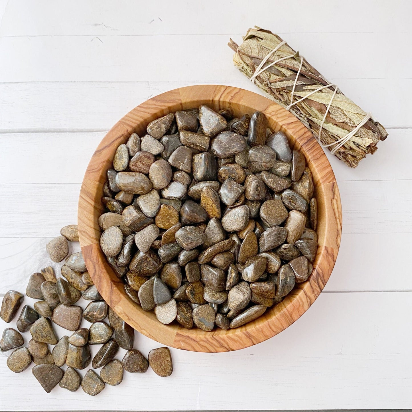 A wooden bowl filled with polished stones, including Bronzite Crystal Chips known for boosting self-confidence, is placed on a white wooden surface. Several stones have spilled out of the bowl onto the surface. To the right of the bowl is a neatly tied bundle of dried herbs.