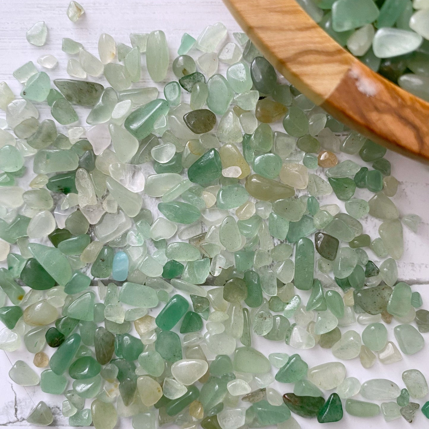 A wooden bowl filled with green and white sea glass pieces sits on a white surface, scattered with tiny Green Aventurine Crystal Chips. Some sea glass pieces are scattered outside the bowl. Small glass bottles with sea glass and a bundle of tied sticks are also present in the background.