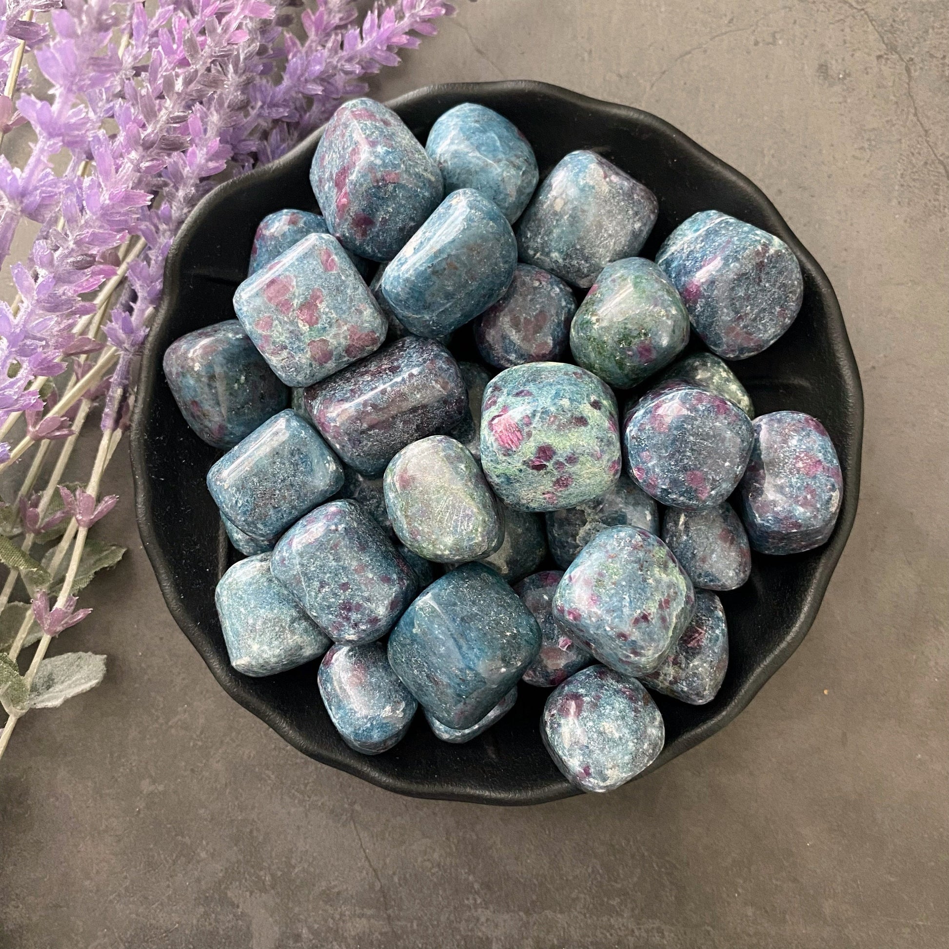 A black bowl filled with shiny, polished Ruby Kyanite Tumbled Stones with pink and green speckles is placed on a dark gray surface. Adjacent to the bowl, on the left side of the image, there are sprigs of purple lavender flowers, invoking high vibrational magick for enhancing psychic abilities.