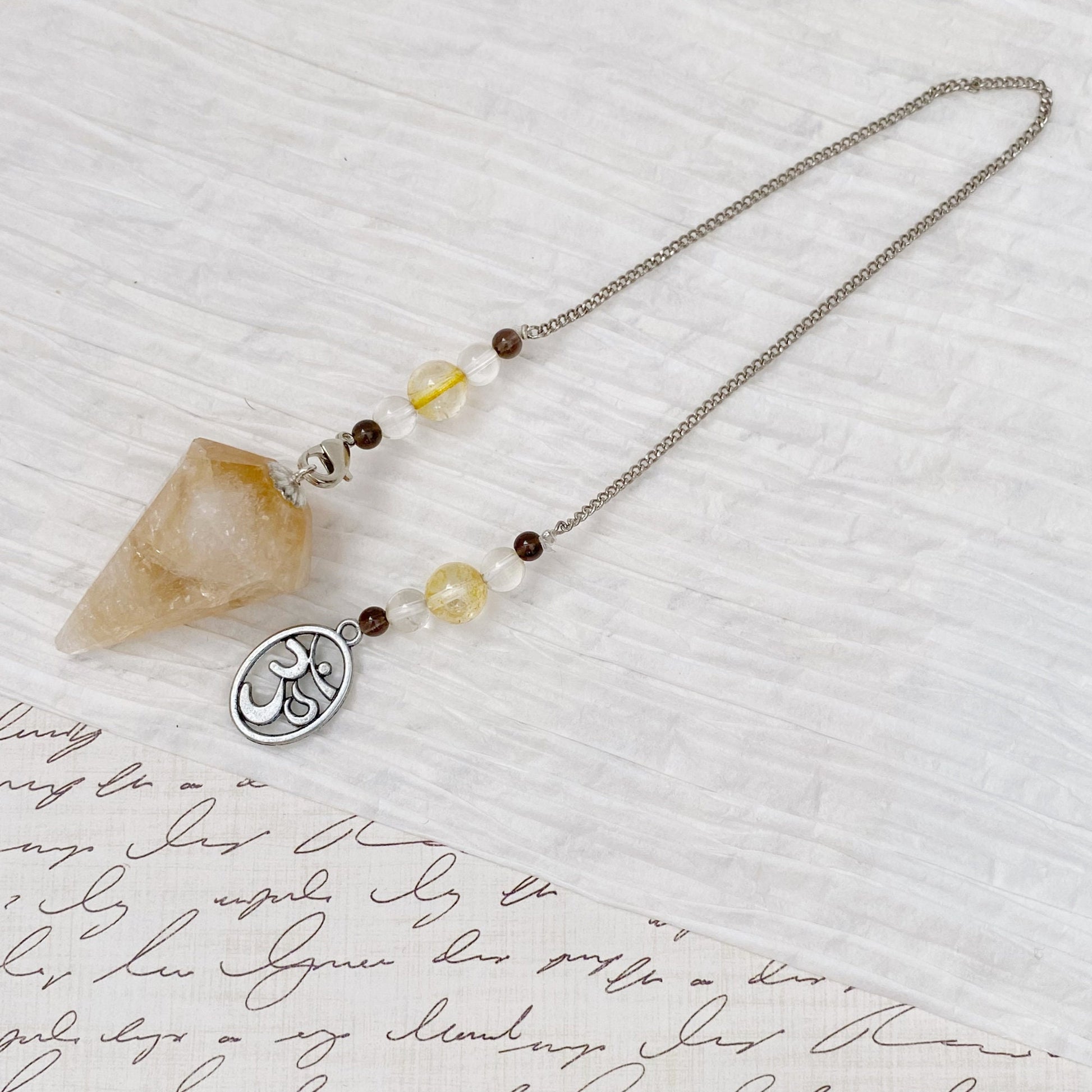 A Citrine Hexagonal Pendulum featuring a pointed, translucent yellow stone is suspended from a chain adorned with beads and a silver Om charm, resting on a textured book cover and white paper background. Ideal for divination practices, it exudes both beauty and spiritual energy.