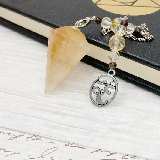 A Citrine Hexagonal Pendulum featuring a pointed, translucent yellow stone is suspended from a chain adorned with beads and a silver Om charm, resting on a textured book cover and white paper background. Ideal for divination practices, it exudes both beauty and spiritual energy.