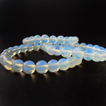 A close-up photograph of an 8mm Opalite Beaded Bracelet against a black background. The spherical, evenly sized beads emit a soft, milky glow with subtle blue and yellow hues. Known as a meditation stone with healing properties, the bracelet's reflection shimmers on the shiny surface below.